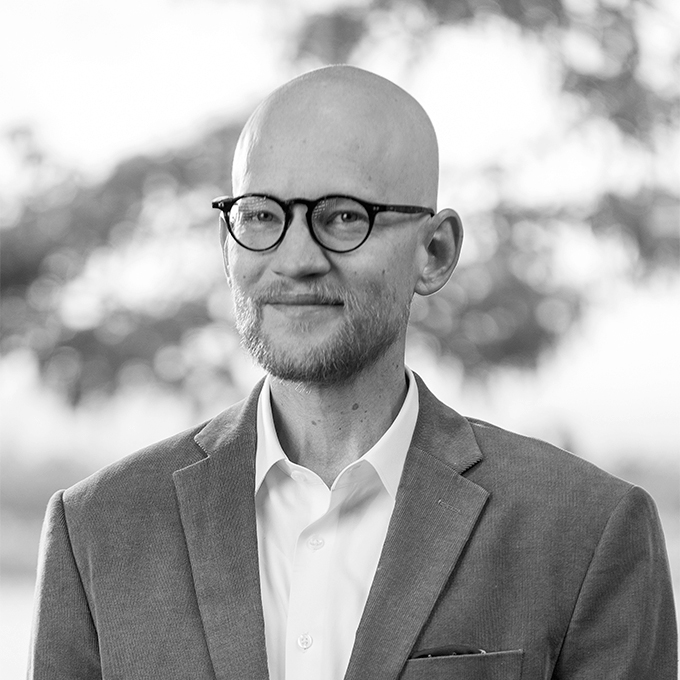 Black and white image of man with glasses in blazer smiling at the camera. This is Tyler Schroeder's bio.