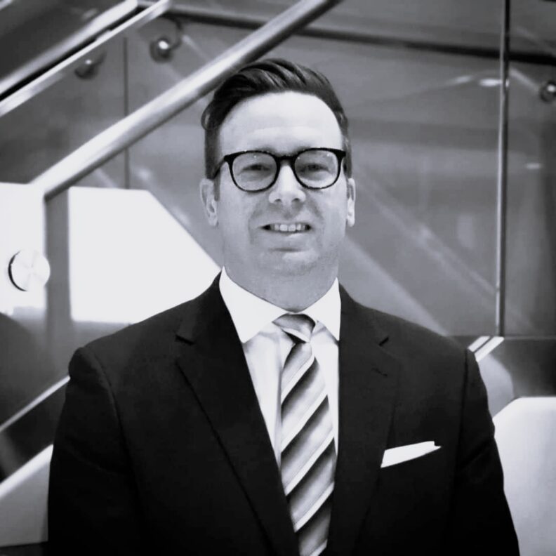 Black and white photo of a man in glasses wearing a suit and tie. This is Scott Devlin's bio